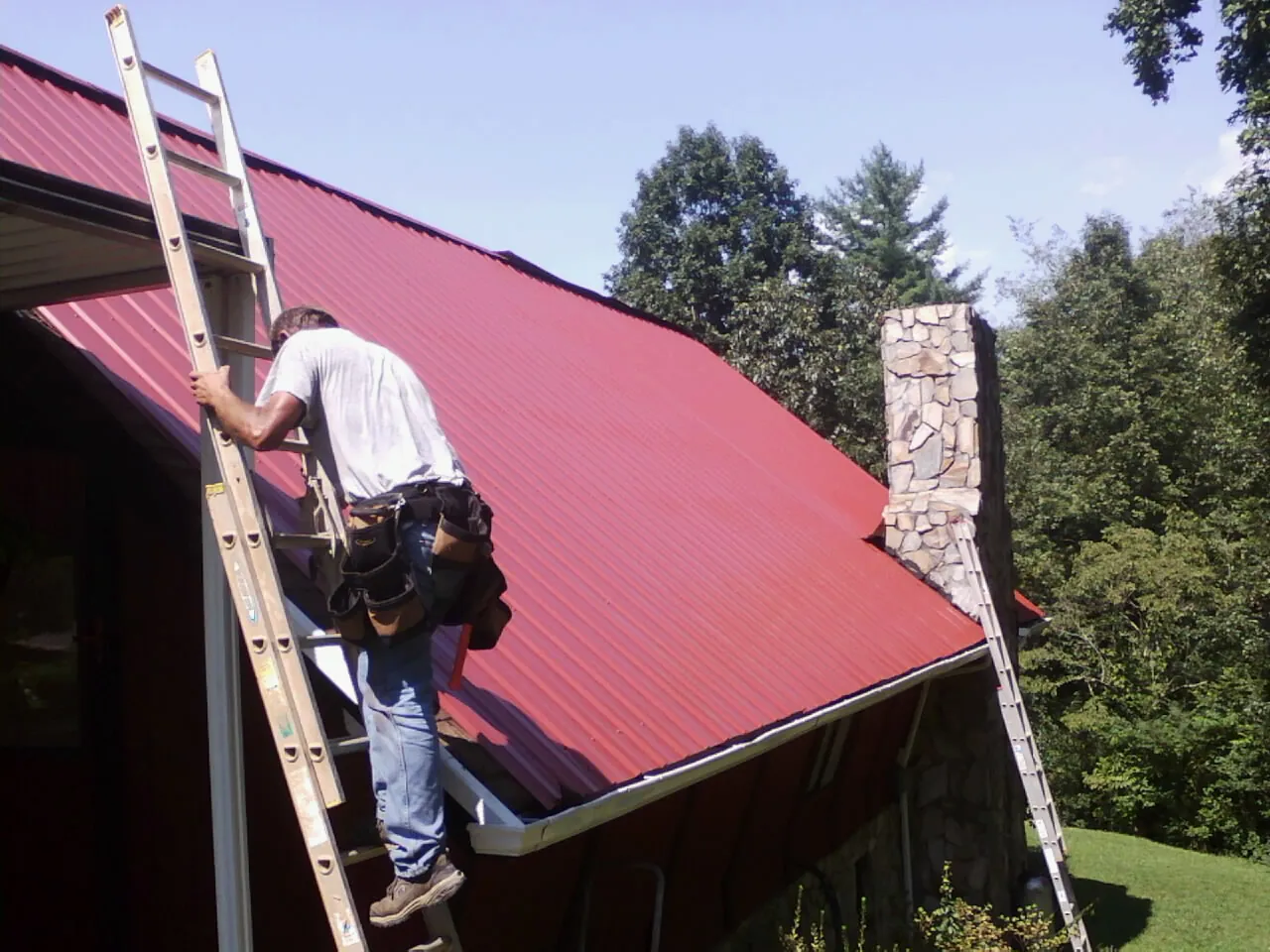 Roofer on a ladder beside a red metal roof and stone chimney — Soffit & Fascia Repair by Roofing Brothers in Elkland